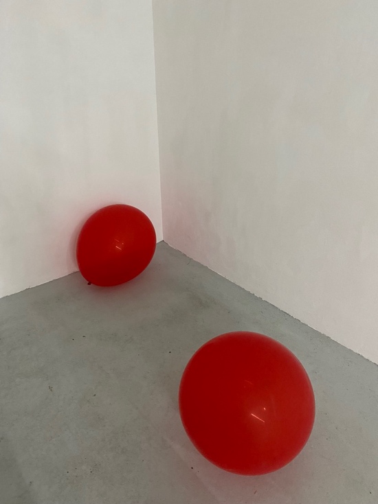 Two red balloons on the floor in the corner of a white gallery